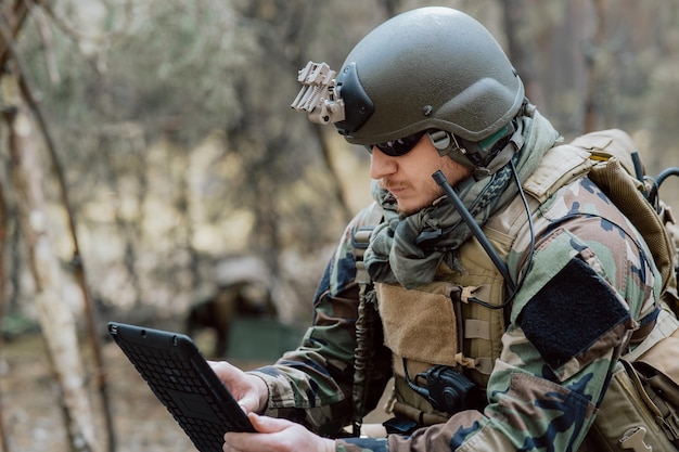 Бородатый солдат в военной форме сидит на военно-транспортных ящиках, анализирует данные на планшете и занимается спортом