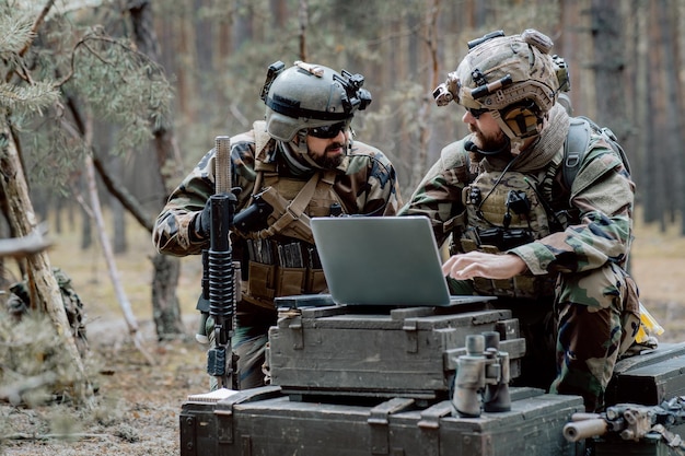 Бородатый солдат в военной форме сидит на военно-транспортных ящиках, анализирует данные на ноутбуке и тренируется