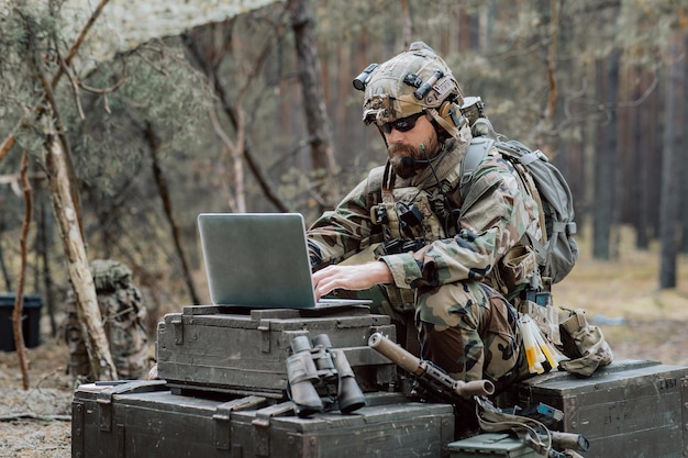 Бородатый солдат в военной форме сидит на военно-транспортных ящиках анализирует данные на ноутбуке и тренируется