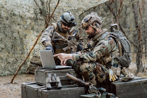 Бородатый солдат в военной форме сидит на военно-транспортных ящиках, анализирует данные на ноутбуке и тренируется